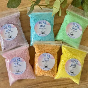 May include: Six small, clear plastic bags filled with colourful, granular bath products labelled "Fizzy Dust." The bags contain pink, purple, blue, green, orange, and yellow contents. Each bag has a circular label with text and a small graphic.