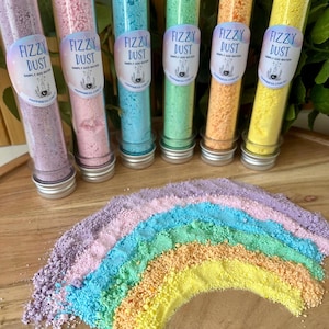May include: Six glass tubes filled with colourful fizzy dust, each with a label that reads "Fizzy Dust". A rainbow of the same coloured dust is spread out below the tubes. The colours include purple, pink, blue, green, orange, and yellow.