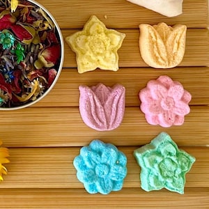 May include: A selection of flower-shaped bath bombs in shades of yellow, pink, blue, and green, presented with a tin of dried flowers and herbs. The bath bombs are displayed on a wooden surface, creating a spa-like atmosphere.