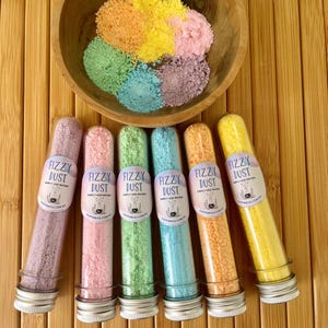 May include: A wooden bowl holds colourful bath bomb powder above six clear tubes of fizzy dust. Each tube contains a different colour of powder, including pink, green, blue, orange, and yellow. The tubes have labels that read "Fizzy Dust".
