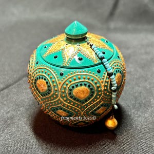 May include: A teal, hand-painted decorative jar with a lid, adorned with gold and orange patterns. The jar features a beaded chain with a gold-colored charm. The text "fragments 2025" is visible on the jar.