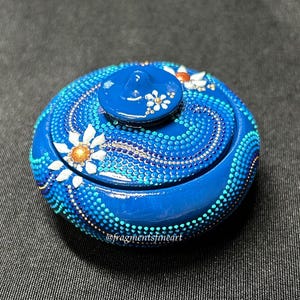 May include: A round blue lidded container decorated with intricate dot art patterns in shades of light blue, dark blue, and gold. The design features painted white flowers with gold centers and the text @fragmentsfineart printed on the side.