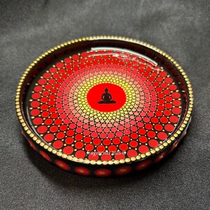 May include: A round, decorative tray with a black rim and a gold beaded edge. The tray features a red and yellow mandala design with a black Buddha silhouette in the center. The text "fragments 2025 ©" is visible.