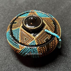May include: A round, black decorative box with a lid, adorned with a geometric pattern of turquoise and orange dots. The lid features a small, black knob and a turquoise beaded handle. The box is a unique, handcrafted item.