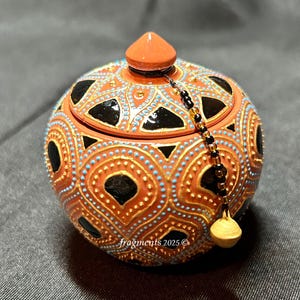Könnte beinhalten: Eine runde, orangefarbene Keramikdose mit Deckel, verziert mit schwarzen, goldenen und blauen Punktmustern. Die Dose hat einen kleinen orangefarbenen Knopf oben und eine Perlenquaste. Der Text "fragments 2025" ist sichtbar.