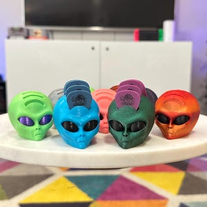 May include: Five alien head guitar pick holders in different colors: green, blue, teal, pink, and orange. Each holder has a slot for guitar picks and the word "Dunlop" printed on the front.