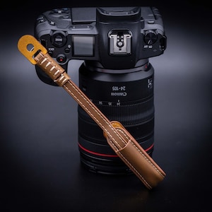 May include: A brown leather camera strap with a gold metal loop. The strap is attached to a black camera lens with the text "Canon 24-105" printed on the side.