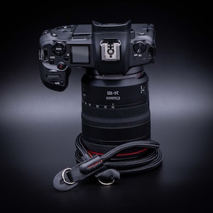 May include: A black Canon camera with a black 24-105 lens attached. The camera is sitting on a black surface with a black camera strap.