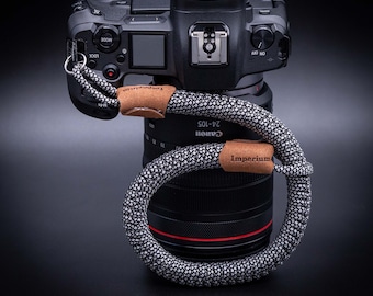 May include: A black camera with a grey and white camera strap with the word "Imperium" on it. The camera lens is a Canon 24-105.