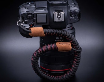 May include: A black Canon camera with a 24-105 lens attached. The camera has a brown leather strap with a black braided cord.