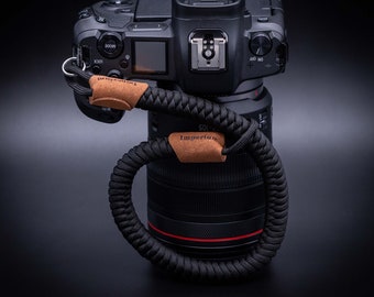 May include: A black camera with a lens attached and a black camera strap with a brown leather patch that says "Imperium" on it.