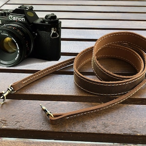 May include: A brown leather camera strap with white stitching and brass clasps. The strap is coiled on a wooden surface next to a black Canon AV-1 camera with a Canon FD 50mm f/1.8 lens.