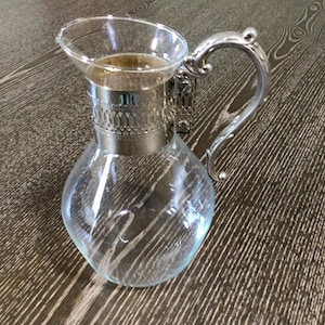 May include: A clear glass pitcher with a silver-colored metal handle and decorative band around the neck. The pitcher has a rounded base and is partially filled with a light-colored liquid. The handle is curved and ornate.