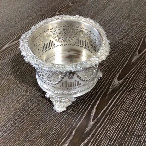 May include: A silver-colored, ornate bowl with intricate cut-out designs and a floral-like border. The bowl has a round shape and sits on three decorative feet. The interior of the bowl is smooth and reflective.