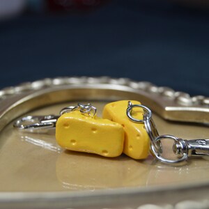 May include: Two small yellow cheese charms attached to a silver key chain. The charms are shaped like wedges of cheese and have a realistic texture.