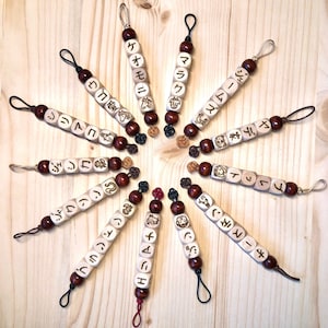 Może przedstawiać: Zestaw 12 drewnianych brelok&oacute;w z japońskimi znakami i symbolami. Każdy brelok ma inny znak i symbol, a wszystkie są nawleczone na czarny sznurek.