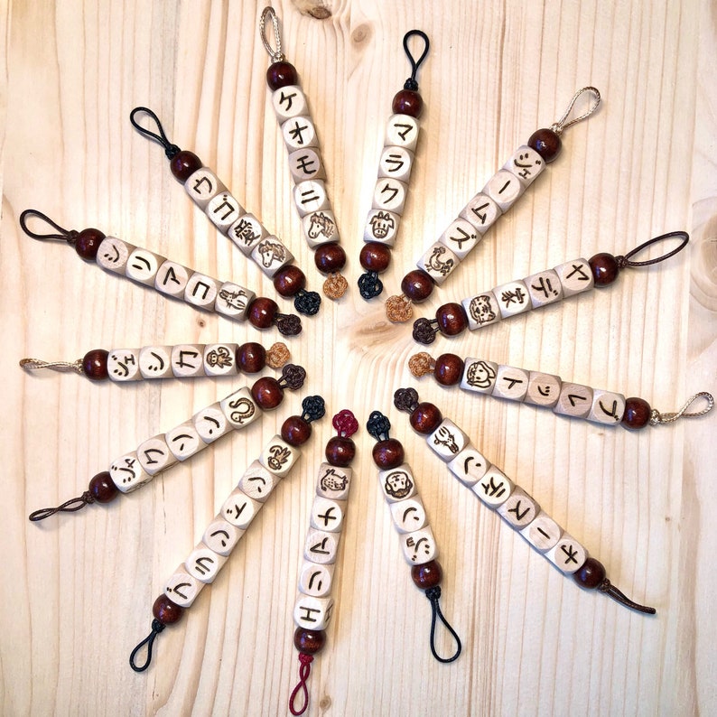 Może przedstawiać: Zestaw 12 drewnianych brelok&oacute;w z japońskimi znakami i symbolami. Każdy brelok ma inny znak i symbol, a wszystkie są nawleczone na czarny sznurek.