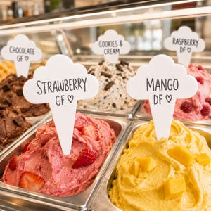 May include: A selection of gelato flavours, including strawberry, mango, and cookies & cream, displayed in metal trays. Each flavour is identified by a white sign. The strawberry gelato is pink, the mango is yellow, and the cookies & cream has chocolate pieces.