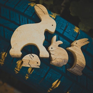 Pu&ograve; includere: Quattro figurine di coniglio in legno, intagliate in uno stile semplice, sono disposte su una superficie testurizzata blu e gialla. I conigli sono di diverse dimensioni e hanno una finitura in legno naturale.