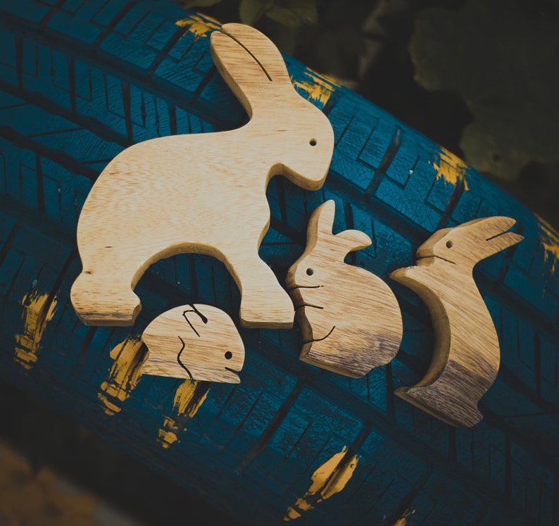 Pu&ograve; includere: Quattro figurine di coniglio in legno, intagliate in uno stile semplice, sono disposte su una superficie testurizzata blu e gialla. I conigli sono di diverse dimensioni e hanno una finitura in legno naturale.