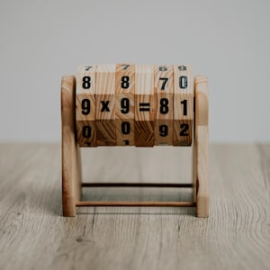 May include: Wooden multiplication learning tool with rotating blocks. The blocks display numbers and the equation 9 x 9 = 81. The wooden frame is light brown, and the blocks have black numbers.
