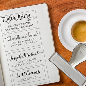 May include: An open notebook with address labels, a coffee cup, and a stamp. The address labels feature names and addresses. The stamp is white and grey. The coffee cup sits on a white saucer. The notebook is open.