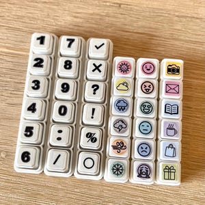 Könnte beinhalten: Eine Sammlung weißer, quadratischer Gummistempel, die in Spalten angeordnet sind. Drei Spalten zeigen schwarz gedruckte Zahlen, mathematische Symbole und Satzzeichen. Zwei Spalten zeigen bunte Symbole, darunter Wettersymbole, Emojis, eine Kamera, einen Umschlag, ein Buch, eine Tasse, eine Tasche und ein Geschenk.