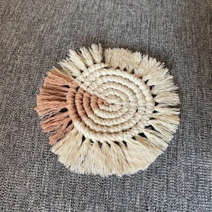 Puede incluir: Un posavasos redondo de macramé blanco y rosa con borde de flecos. El posavasos está hecho con un patrón circular de cuerdas anudadas.