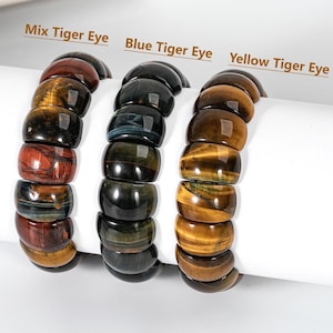 May include: Three beaded bracelets made of tiger eye stones. The bracelets are labeled "Mix Tiger Eye", "Blue Tiger Eye", and "Yellow Tiger Eye". The bracelets are displayed on a white cylindrical stand.