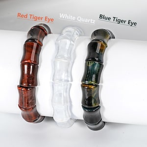 May include: Three beaded bracelets with bamboo-shaped beads. The bracelets are labelled "Red Tiger Eye", "White Quartz", and "Blue Tiger Eye". The Red Tiger Eye bracelet is a dark reddish-brown, the White Quartz is clear, and the Blue Tiger Eye is dark with blue and gold hues.