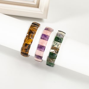 Puede incluir: Tres pulseras de piedras preciosas con cuentas rectangulares en ojo de tigre, amatista y cuarzo rosa, y ágata musgosa verde. Las pulseras se exhiben sobre una superficie blanca. Un marco de fotos blanco está en el fondo.