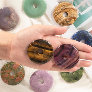 Puede incluir: Surtido de colgantes de rosquilla de piedras preciosas pulidas en varios colores, incluyendo verde, morado, marrón y azul. Cada colgante tiene un agujero central. Los colgantes se muestran sobre una superficie blanca.