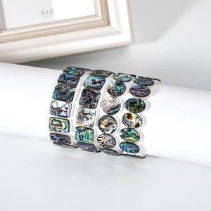 May include: A stack of four bracelets with iridescent abalone shell beads. The bracelets feature square and oval-shaped beads in shades of blue, green, and purple. The bracelets are displayed on a white cylindrical surface.