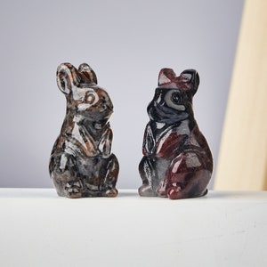 May include: Two carved stone rabbit figurines. The rabbits are in a seated position with their ears upright. One rabbit is primarily brown and gray, while the other is dark red and black. The figurines are on a white surface.