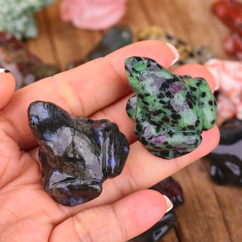 K&ouml;nnte beinhalten: Zwei geschnitzte Froschfiguren, eine gr&uuml;n mit schwarzen Flecken und die andere schwarz mit blauen Sprenkeln. Die Figuren sind aus poliertem Stein gefertigt und werden auf einer Hand pr&auml;sentiert.