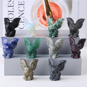 Peut inclure: Collection de petites figurines de gargouilles en pierre sculptée de différentes couleurs, dont noir, blanc, vert, bleu et gris. Chaque figurine a des ailes détaillées et une pose assise. Les figurines sont exposées sur une surface grise.