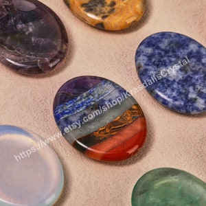 Op de afbeelding: Zeven chakra stenen in verschillende kleuren, waaronder paars, blauw, groen, geel, oranje en rood, gerangschikt in een rij op een lichtbruin oppervlak. De stenen zijn glad en ovaalvormig. De tekst "https://www.etsy.com/shop/KrasdallsCrystal" is zichtbaar in de afbeelding.