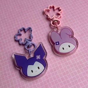Könnte beinhalten: Zwei Schlüsselanhänger aus Acryl mit Cartoon-Charakter-Designs. Einer ist lila mit einem katzenartigen Gesicht, der andere rosa mit einem hasenartigen Gesicht. Beide haben passende blütenförmige Verschlüsse auf rosa Hintergrund.