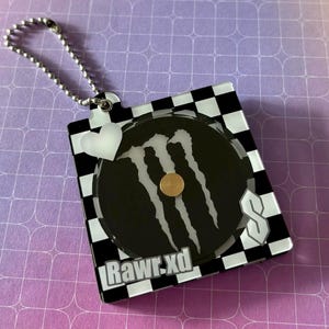 Könnte beinhalten: Ein Schlüsselanhänger mit einem schwarz-weiß karierten Rand. In der Mitte befindet sich ein schwarzer Kreis mit einem weißen Monster Energy-Logo und einem goldfarbenen Kreis in der Mitte. Der Text "Rawr.xd" befindet sich unten.