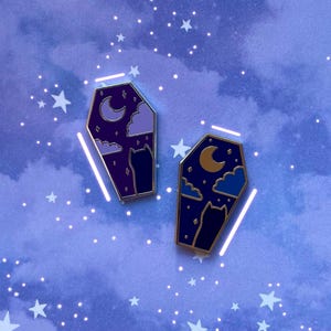 May include: Two coffin-shaped enamel pins with gold edging. One pin has a purple background with a crescent moon, clouds, and a cat silhouette. The other pin has a navy blue background with a gold moon, clouds, and a cat silhouette.