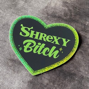 Könnte beinhalten: Ein herzförmiger Aufkleber mit dem Text "SHREXY Bitch" in leuchtendem Grün, umrandet von einem glitzernden, irisierenden Rand. Der Hintergrund des Aufklebers ist schwarz. Der Aufkleber befindet sich auf einer grauen Stoffoberfläche.