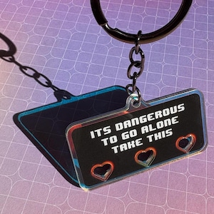 Könnte beinhalten: Ein rechteckiger Schlüsselanhänger mit dem Text "ITS DANGEROUS TO GO ALONE TAKE THIS" in Weiß auf schwarzem Hintergrund. Der Schlüsselanhänger hat drei herzförmige Aussparungen und ist an einem schwarzen Schlüsselring befestigt.