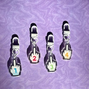May include: Four clear acrylic keyrings shaped like a hand giving the middle finger, each with a different number from 1 to 4. The numbers are in different colours. Each keyring has a silver clasp.
