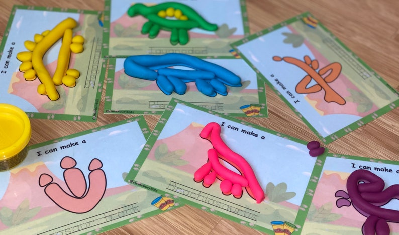 K&ouml;nnte beinhalten: Sechs Karten mit Dinosaurierumrissen und dem Text "Ich kann ein" gefolgt vom Namen des Dinosauriers. Jede Karte hat einen Dinosaurier aus Knetmasse in einer anderen Farbe, der an den Umriss angepasst ist.