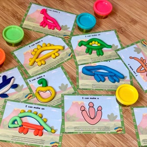 K&ouml;nnte beinhalten: Ein Set mit 10 druckbaren Dinosaurier-Knetmatten mit verschiedenen Dinosaurierformen. Jede Matte enth&auml;lt den Text "Ich kann ein" oder "Ich kann eine" gefolgt vom Namen des Dinosauriers. Die Matten sind auf wei&szlig;em Karton gedruckt und haben einen gr&uuml;nen Rand. Es gibt auch mehrere Beh&auml;lter mit Knetmasse in verschiedenen Farben.