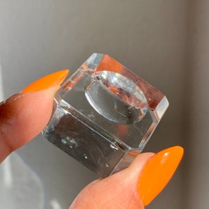 May include: A clear glass ring holder with a square base. The holder is designed to display a ring.