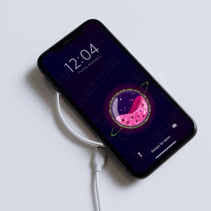 Könnte beinhalten: Ein schwarzes Smartphone mit einer Wassermelonen-Tapete wird auf einem weißen kabellosen Ladegerät geladen. Der Bildschirm des Telefons zeigt die Uhrzeit, das Datum und den Text "CLUB LORE".