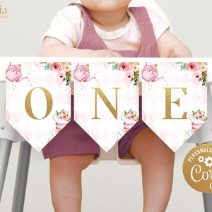 May include: A pink and gold high chair banner with the word "ONE" in gold letters. The banner features a teacup and teapot design with floral accents. The banner is decorated with gold glitter and pink and white diamond patterns.
