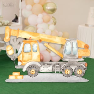 May include: A yellow and gray toy construction crane with a long arm and a hook. The crane is on a green surface with a few yellow blocks in front of it.