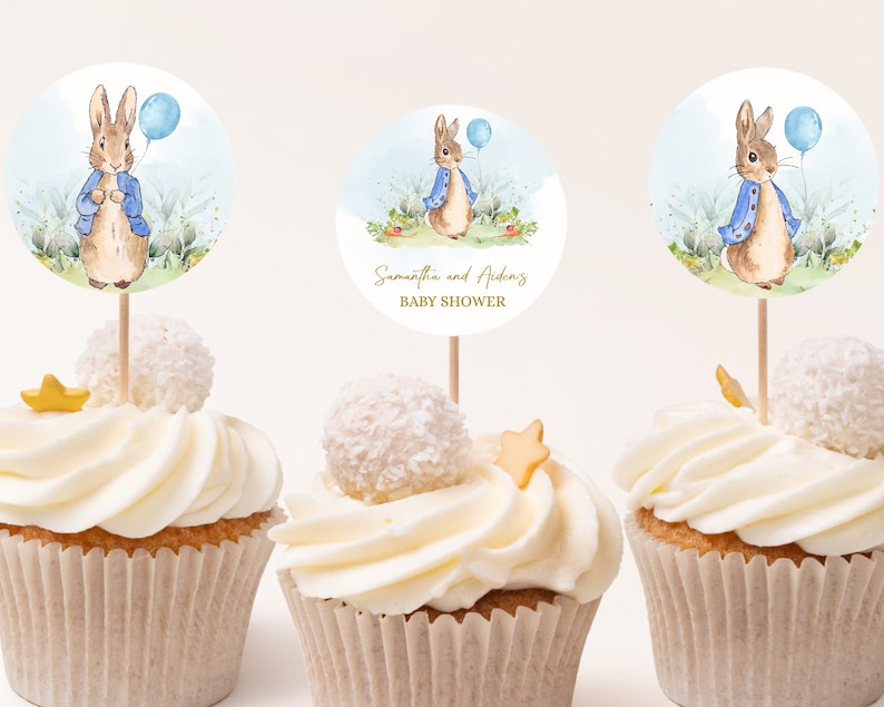 May include: Three cupcakes decorated with white frosting and a blue and brown Peter Rabbit themed topper. The topper features a rabbit holding a blue balloon and the text "Samantha and Aiden's Baby Shower".